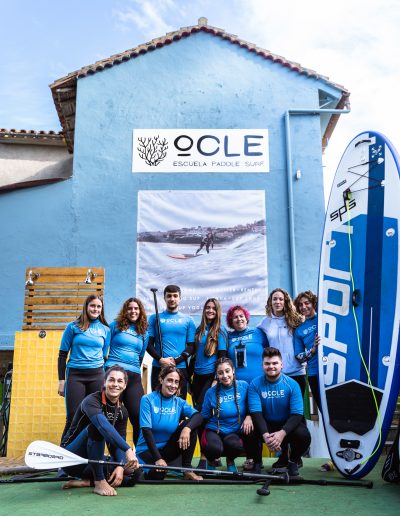 Un grupo de personas vestidas con trajes azules iguales posan con remos y una tabla de remo frente a un edificio con un cartel de "Escuela Paddle Surf".