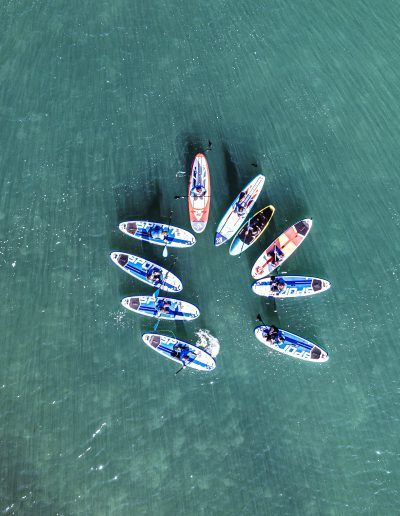 Vista aérea de ocho personas remando en tablas de surf de remo en formación circular sobre aguas verdes claras.