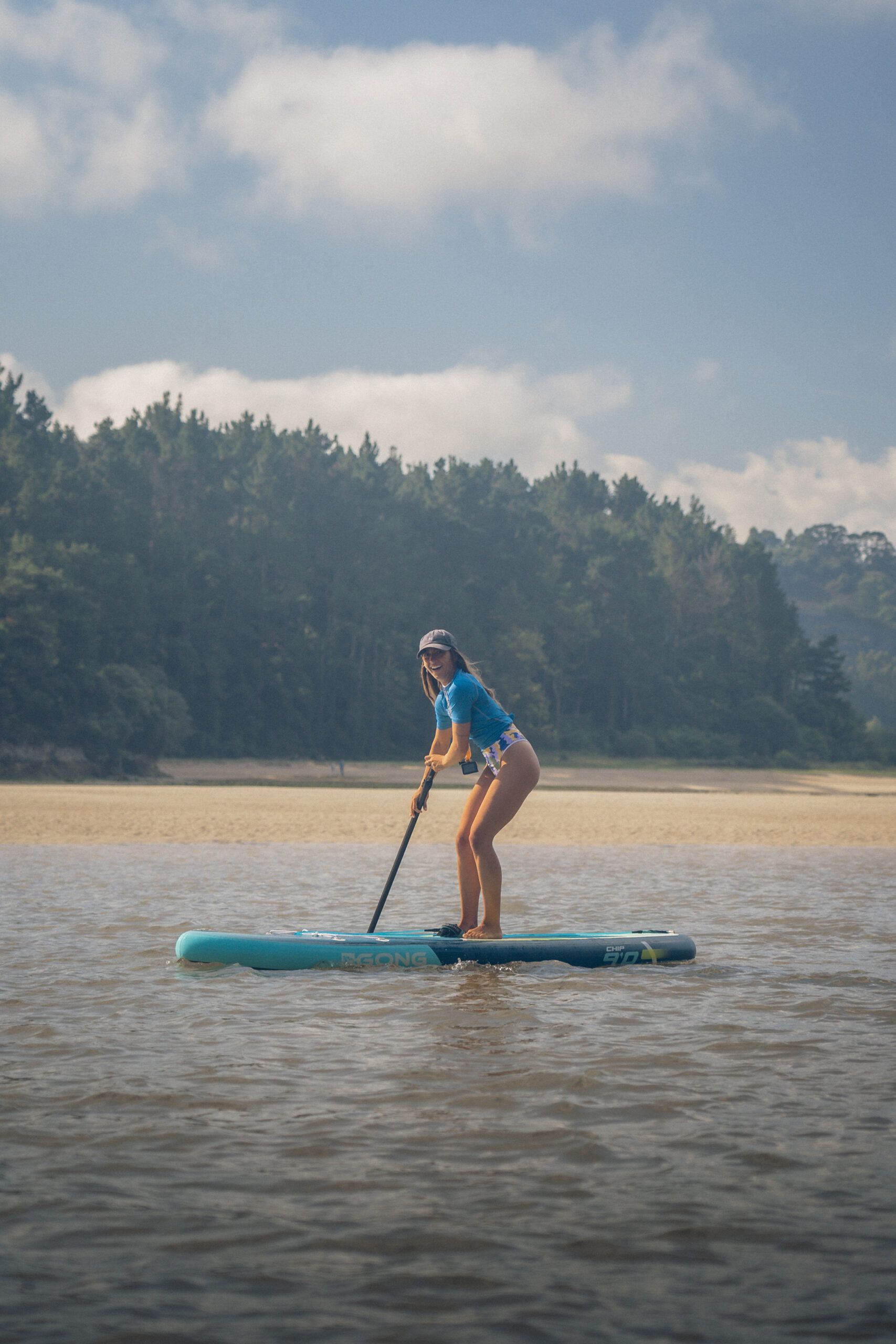 Ocle-014 Persona haciendo paddleboard en aguas tranquilas con playa de arena y bosque al fondo.