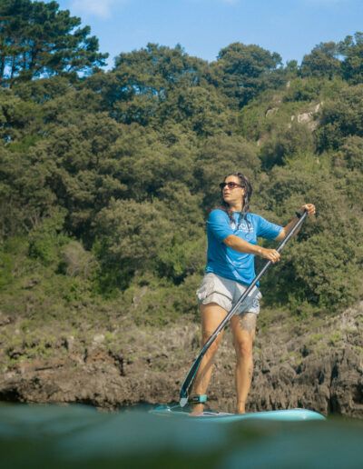 Persona practicando paddle surf en aguas tranquilas cerca de una costa rocosa y arbolada. Clima soleado. Viste una camisa azul y pantalones cortos grises.