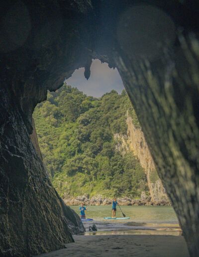 Vista desde el interior de una cueva rocosa hacia dos personas haciendo paddleboard en un mar en calma, con exuberantes colinas verdes en el fondo.